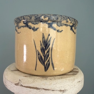 May include: A cylindrical ceramic crock with a tan base and a blue sponge-painted rim. The front features a hand-painted blue wheat stalk design centered between two vertical lines. The vessel has a smooth, glossy finish and a wide, open top.