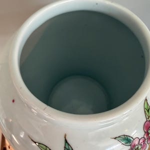 Pode incluir: Um pote de cer&acirc;mica branca com uma boca larga e aberta e um interior liso em azul claro. O exterior apresenta motivos bot&acirc;nicos pintados &agrave; m&atilde;o, incluindo flores rosa vibrantes e folhas verdes delineadas com tinta escura, sobre um acabamento brilhante.