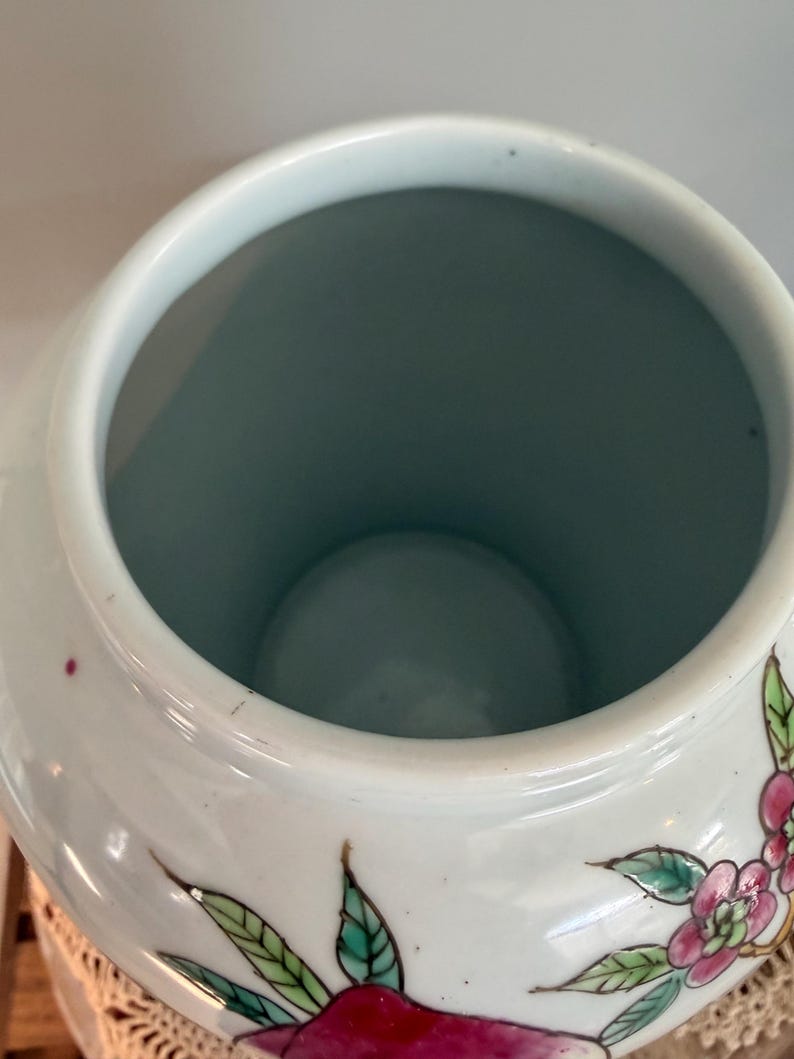 Pode incluir: Um pote de cer&acirc;mica branca com uma boca larga e aberta e um interior liso em azul claro. O exterior apresenta motivos bot&acirc;nicos pintados &agrave; m&atilde;o, incluindo flores rosa vibrantes e folhas verdes delineadas com tinta escura, sobre um acabamento brilhante.