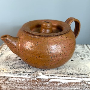 May include: A handcrafted, round brown ceramic teapot with a lid and handle. The teapot has a spout and a textured surface with dark speckles. The teapot is sitting on a white and gray wooden surface.