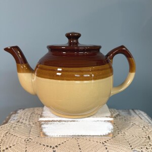 May include: A ceramic teapot featuring a tri-color design with a dark brown lid and upper section, a medium brown middle band, and a light beige base. The teapot includes a curved spout, a matching handle, and a removable lid with a knob handle.