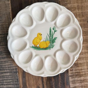 May include: A round, off-white ceramic egg plate featuring a scalloped edge and twelve circular indentations for holding eggs. The center of the plate displays a colorful illustration of two yellow chicks standing near green grass and tall stalks.