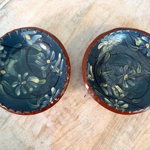May include: Two round ceramic bowls with a dark blue interior featuring hand-painted light yellow floral patterns and thin black swirling lines. The bowls have a smooth, glossy finish and a distinct reddish-brown rim and exterior base.