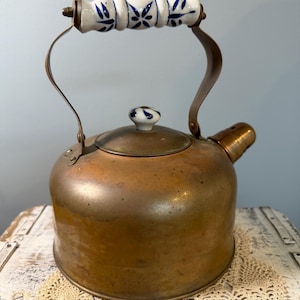 May include: Vintage copper kettle with a white and blue floral patterned handle and lid knob. The kettle has a rounded body, a spout, and a curved metal handle. The kettle is sitting on a white surface with a beige lace doily.