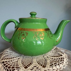 May include: A green ceramic teapot with a gold band and floral design. The teapot has a curved handle, a spout, and a lid with a small knob. The teapot is sitting on a white lace doily.