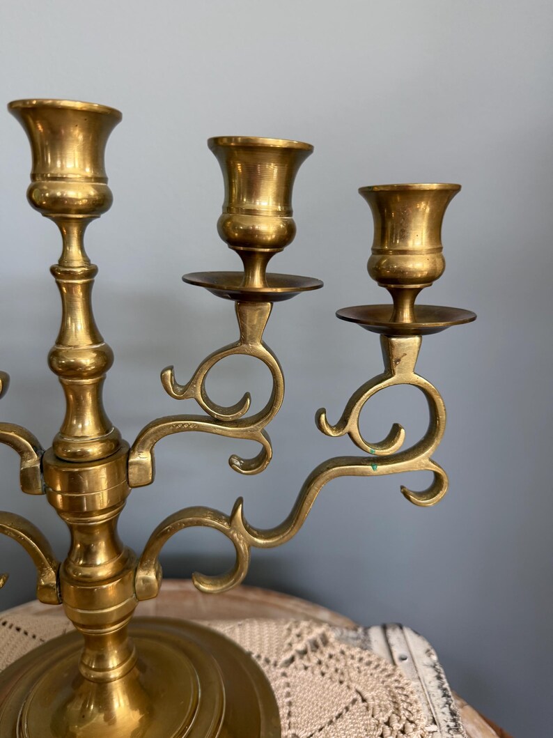 Puede incluir: Un candelabro de lat&oacute;n con tres candeleros. El candelabro tiene una base redonda y brazos curvos. Los candeleros tienen forma de copas. El candelabro es de color dorado c&aacute;lido y tiene un aspecto antiguo.