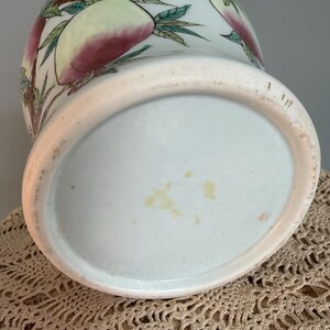 Pode incluir: Um vaso de cer&acirc;mica com uma cor base azul-esverdeada p&aacute;lida, apresentando um design pintado &agrave; m&atilde;o de ramos de pessegueiro com folhas verdes e p&ecirc;ssegos grandes e arredondados em tons de rosa e amarelo. A base circular n&atilde;o &eacute; esmaltada.