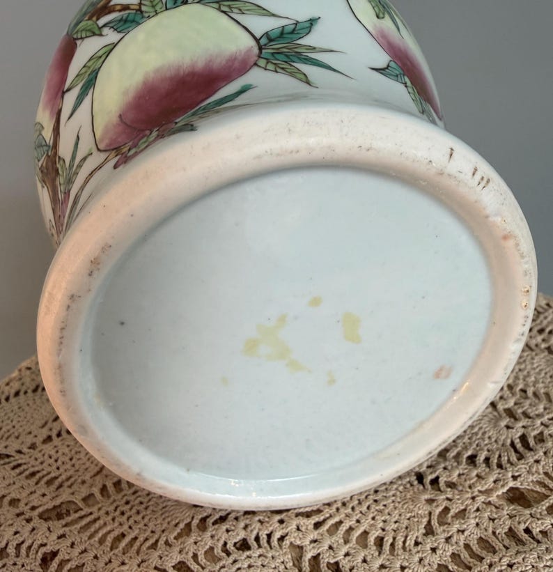 Pode incluir: Um vaso de cer&acirc;mica com uma cor base azul-esverdeada p&aacute;lida, apresentando um design pintado &agrave; m&atilde;o de ramos de pessegueiro com folhas verdes e p&ecirc;ssegos grandes e arredondados em tons de rosa e amarelo. A base circular n&atilde;o &eacute; esmaltada.