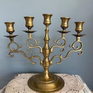 Puede incluir: Un candelabro de lat&oacute;n vintage con cinco candeleros. El candelabro tiene una columna central y brazos curvos, cada uno con una copa para velas. La base es redonda y el dise&ntilde;o general es ornamentado y decorativo. El candelabro es de lat&oacute;n.