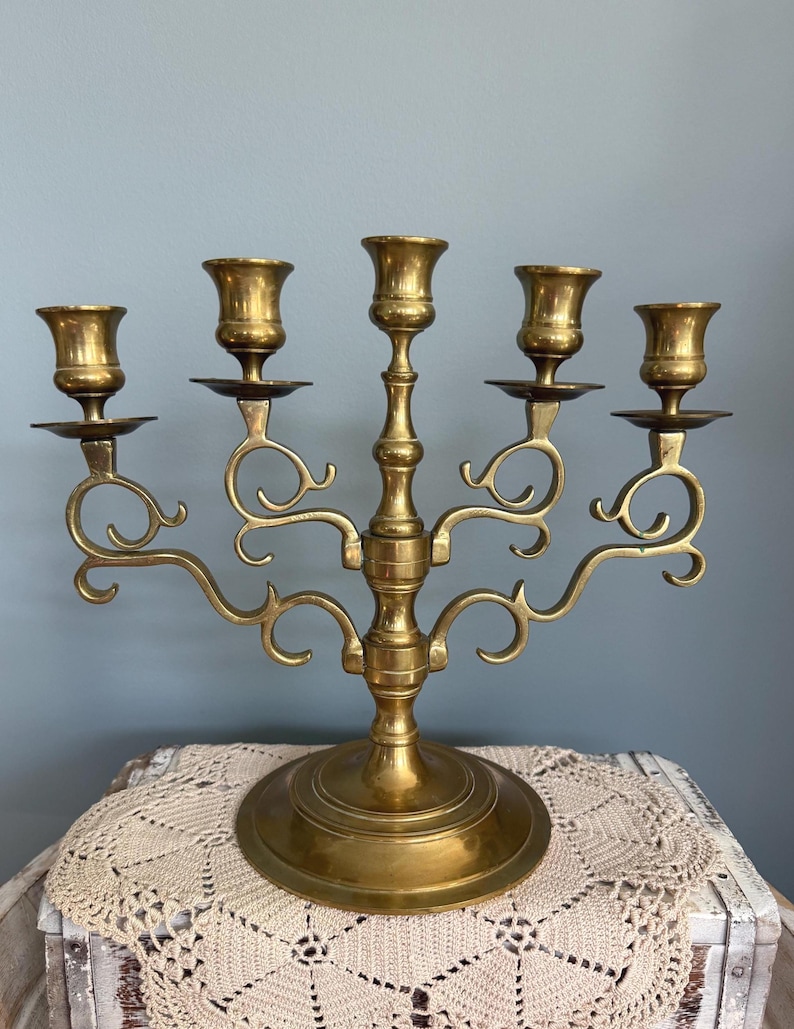 Puede incluir: Un candelabro de lat&oacute;n vintage con cinco candeleros. El candelabro tiene una columna central y brazos curvos, cada uno con una copa para velas. La base es redonda y el dise&ntilde;o general es ornamentado y decorativo. El candelabro es de lat&oacute;n.
