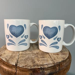 May include: Two white ceramic mugs with blue heart designs. Each mug features a large heart in the center and smaller hearts and decorative elements around it. The mugs have a handle and are displayed on a wooden surface.