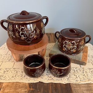 Puede incluir: Un juego de cuatro fuentes de cerámica marrón con tapas y asas. Los platos presentan diseños florales en blanco, naranja y dorado. El juego incluye dos ollas con tapa y dos cuencos más pequeños, ideales para servir sopas o guisos.