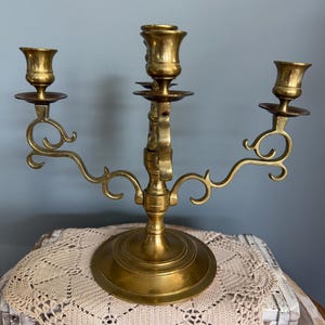 Puede incluir: Un candelabro de lat&oacute;n con tres candeleros. El candelabro tiene una base redonda y brazos curvos con adornos decorativos. Los candeleros tienen forma de peque&ntilde;as copas. El candelabro se exhibe sobre un mantel individual de ganchillo color crema.