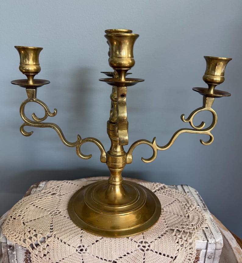Puede incluir: Un candelabro de lat&oacute;n con tres candeleros. El candelabro tiene una base redonda y brazos curvos con adornos decorativos. Los candeleros tienen forma de peque&ntilde;as copas. El candelabro se exhibe sobre un mantel individual de ganchillo color crema.