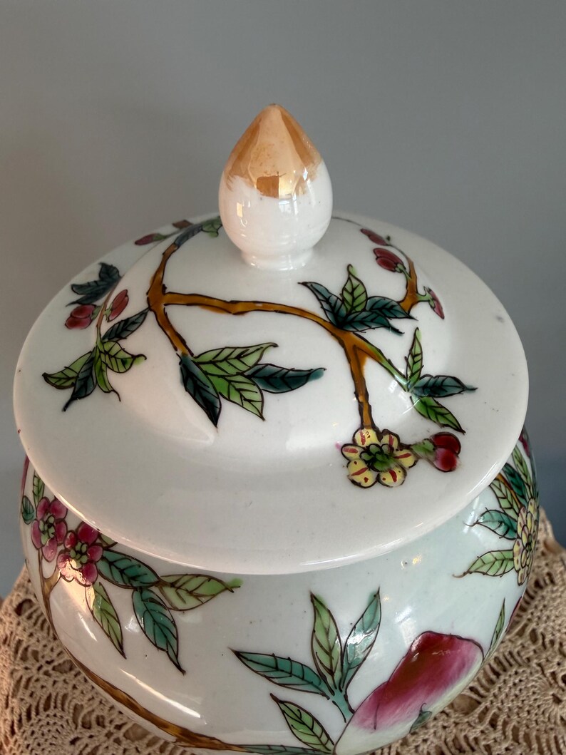 Pode incluir: Um pote de cer&acirc;mica branca com tampa correspondente e um remate pintado a ouro. A superf&iacute;cie &eacute; decorada com ilustra&ccedil;&otilde;es pintadas &agrave; m&atilde;o de ramos castanhos, folhas verdes e flores cor-de-rosa e amarelas. O pote tem uma forma arredondada.