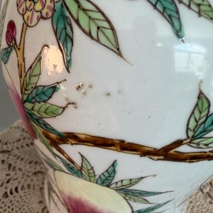 Pode incluir: Um vaso de cer&acirc;mica branca com um design pintado &agrave; m&atilde;o de ramos castanhos, folhas verdes e flores cor-de-rosa e amarelas. A superf&iacute;cie apresenta um acabamento brilhante com pinceladas vis&iacute;veis e pequenas manchas escuras no fundo.