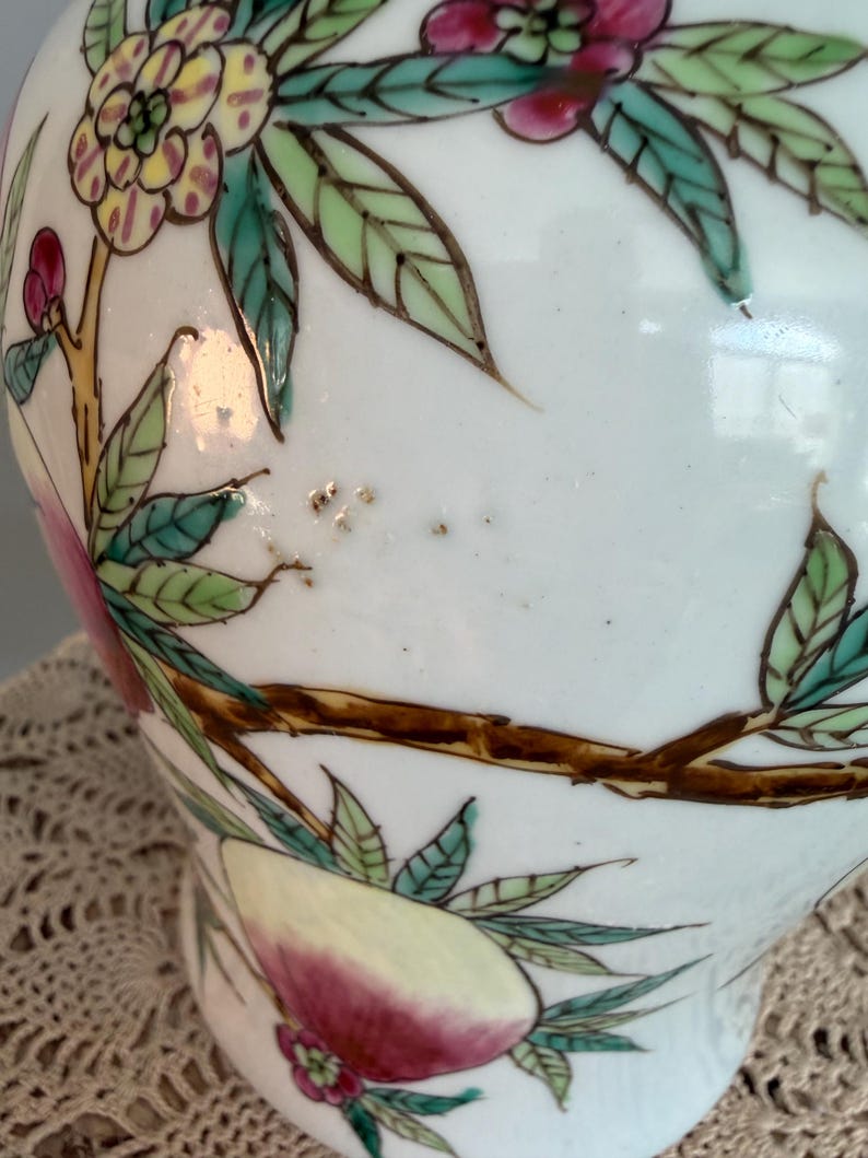 Pode incluir: Um vaso de cer&acirc;mica branca com um design pintado &agrave; m&atilde;o de ramos castanhos, folhas verdes e flores cor-de-rosa e amarelas. A superf&iacute;cie apresenta um acabamento brilhante com pinceladas vis&iacute;veis e pequenas manchas escuras no fundo.