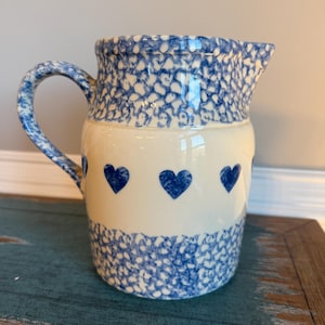 Puede incluir: Jarra de cerámica con diseño azul y blanco. La parte superior e inferior de la jarra tienen un patrón moteado azul, mientras que el centro presenta tres formas de corazón azul. El asa también es azul y blanca.