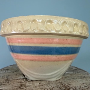 May include: A cream-colored ceramic mixing bowl featuring a wide, scalloped rim and horizontal bands of pink and blue glaze around the center. The exterior surface displays a smooth, glossy finish with subtle molded detailing near the base.