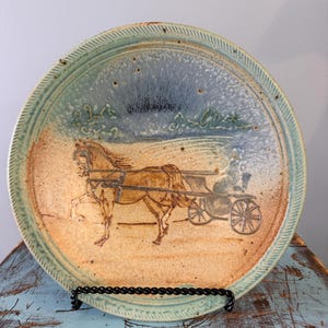 May include: A round ceramic plate featuring a hand-drawn illustration of a horse pulling a carriage. The plate has a speckled tan base with a blue-toned upper section and a textured, ridged rim. The horse and carriage are rendered in brown.