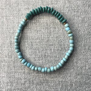May include: A beaded bracelet featuring turquoise and teal beads. The bracelet is made of small, round beads in varying shades of blue and green, strung together with a gold-colored accent. The bracelet is designed to be worn around the wrist.
