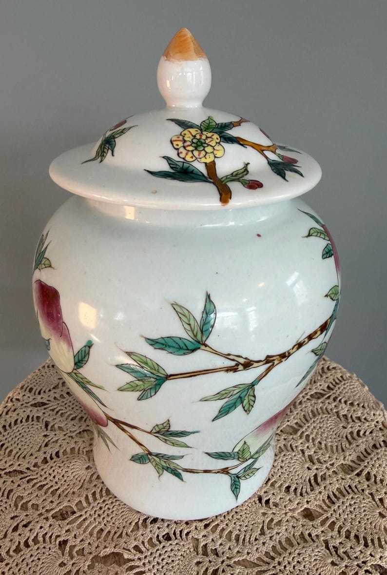 Pode incluir: Um pote de gengibre de cer&acirc;mica branca com tampa correspondente e um remate pontiagudo. O pote &eacute; decorado com ilustra&ccedil;&otilde;es pintadas &agrave; m&atilde;o de ramos verdes com folhas, flores amarelas e frutos vermelho-rosados num estilo delicado.