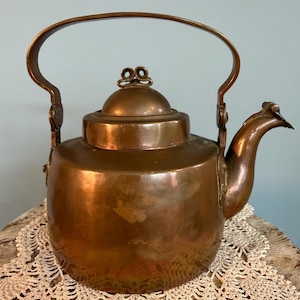 May include: A copper kettle with a large arched handle and a spout. The kettle has a rounded body and a lid with a decorative knob. The copper has a warm, reddish-brown color, and the kettle appears to be vintage.