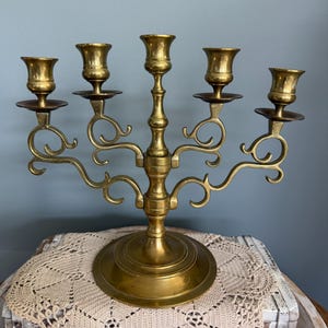 Puede incluir: Un candelabro de lat&oacute;n con cinco candeleros. El candelabro tiene una base redonda y brazos curvos. Los candeleros tienen forma de peque&ntilde;as copas. El candelabro est&aacute; sobre un mantelito color crema.