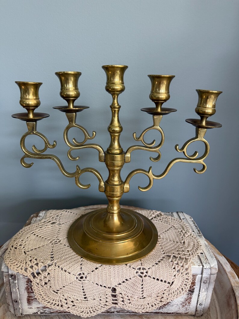 Puede incluir: Un candelabro de lat&oacute;n con cinco candeleros. El candelabro tiene una base redonda y brazos curvos. Los candeleros tienen forma de peque&ntilde;as copas. El candelabro est&aacute; sobre un mantelito color crema.