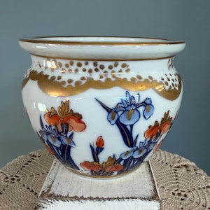 May include: A white ceramic planter with a gold rim and decorative gold accents. The planter features a floral design with orange and blue irises. The planter is round with a wide opening.