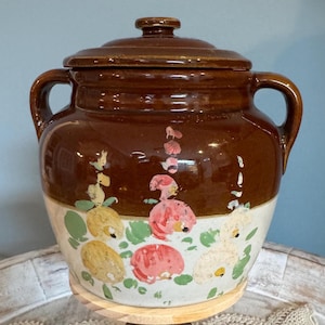 May include: A two-toned ceramic crock with a lid and two side handles. The upper portion is a glossy dark brown, while the lower section is a cream color featuring hand-painted floral motifs in yellow, pink, and green with small green leaves.