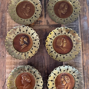May include: Six antique bronze-colored medallions featuring portraits of historical figures. Each medallion is set within a decorative, gold-colored, floral-patterned frame. The medallions are arranged on a rustic, wooden surface.