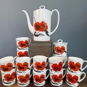 Peut inclure: Service à thé en céramique blanche avec un motif de coquelicots rouges. L'ensemble comprend une théière haute, un sucrier avec couvercle et plusieurs tasses. Le motif de coquelicots est répété sur chaque pièce.