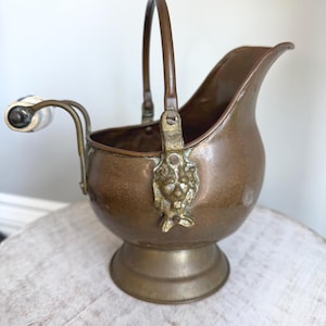 May include: A vintage copper coal scuttle with a brass handle and decorative lion's head accents. The scuttle has a curved spout and a white and black handle. The base is a pedestal design.