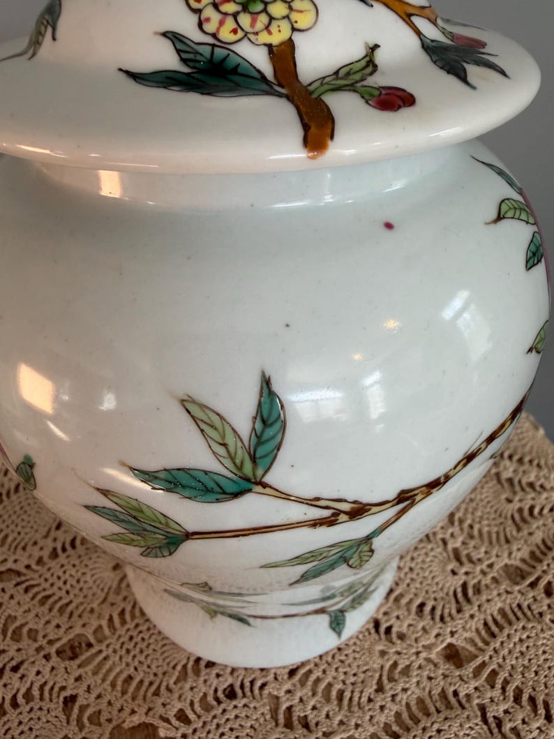 Pode incluir: Um pote de gengibre de cer&acirc;mica branca com tampa correspondente, apresentando ilustra&ccedil;&otilde;es bot&acirc;nicas pintadas &agrave; m&atilde;o de ramos castanhos, folhas verdes e flores amarelas e vermelhas. O pote tem um corpo arredondado e uma base alargada.