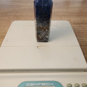 May include: A polished sodalite crystal obelisk, with a deep blue colour and speckled patterns of grey, brown, and white. The obelisk is on a white scale, weighing 96 grams. The top of the obelisk is a pointed pyramid shape.