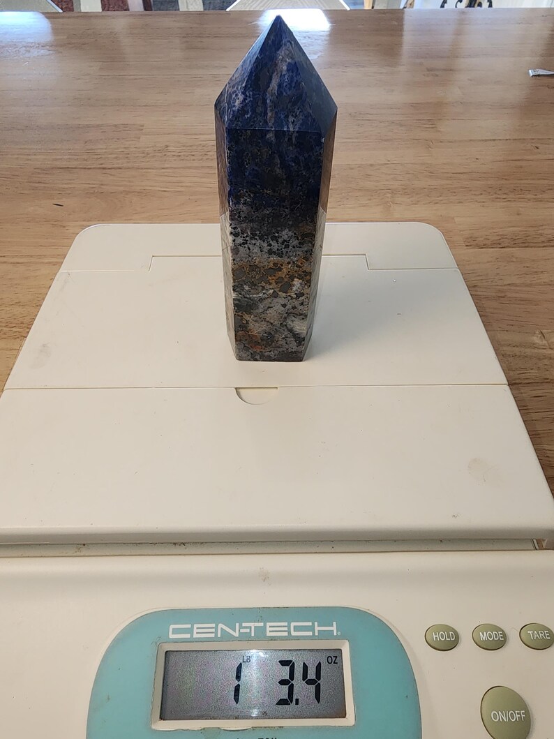 May include: A polished sodalite crystal obelisk, with a deep blue colour and speckled patterns of grey, brown, and white. The obelisk is on a white scale, weighing 96 grams. The top of the obelisk is a pointed pyramid shape.