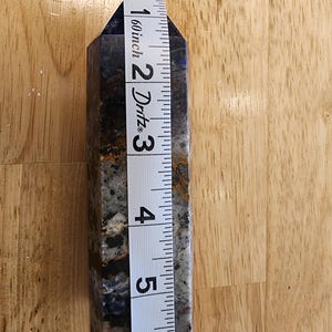 May include: A blue and grey crystal obelisk, approximately 15 cm tall, is measured with a white tape measure. The obelisk has a polished, faceted surface with dark blue and grey marbling and some brown inclusions.