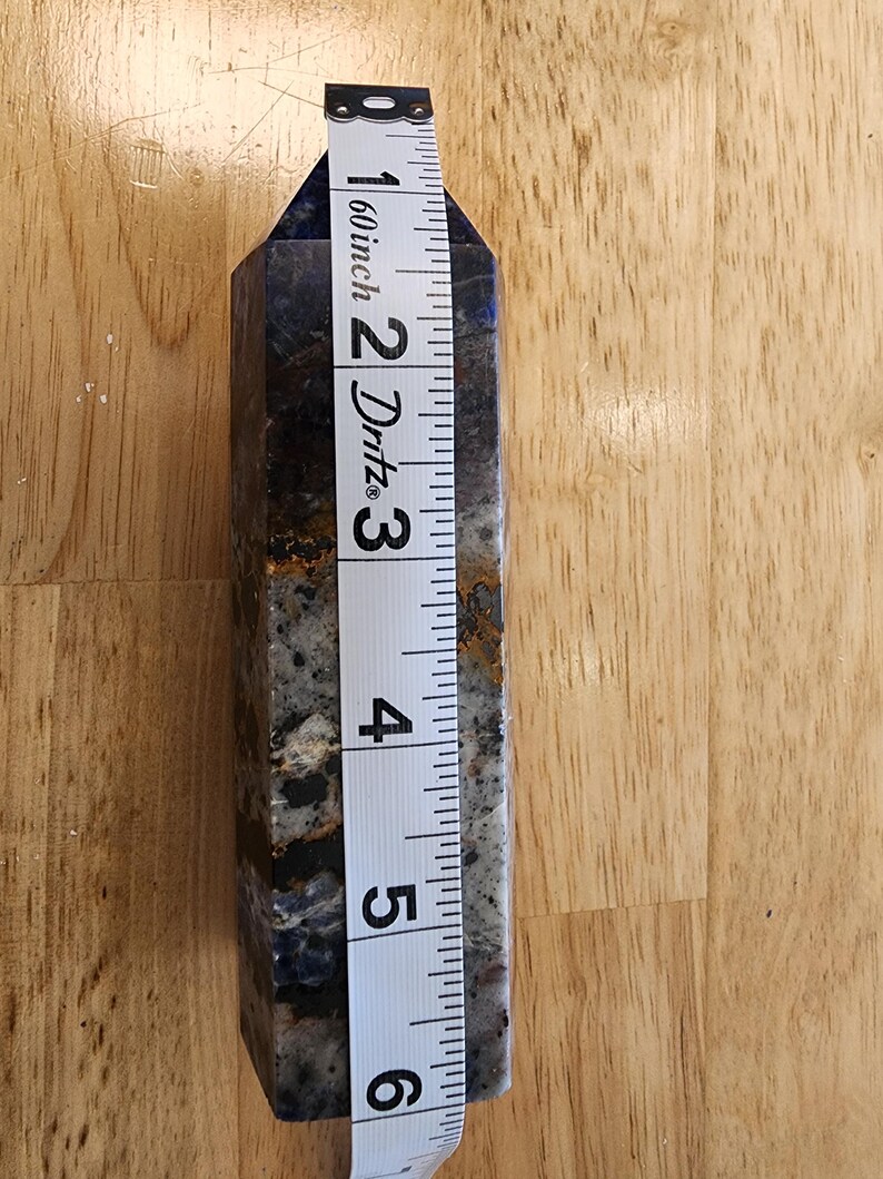 May include: A blue and grey crystal obelisk, approximately 15 cm tall, is measured with a white tape measure. The obelisk has a polished, faceted surface with dark blue and grey marbling and some brown inclusions.