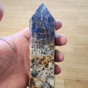 May include: A rectangular crystal point with a flat base and a pointed top. The crystal features a mix of colours, including deep blue, white, grey, and orange, with a polished surface. The crystal is held in a hand.