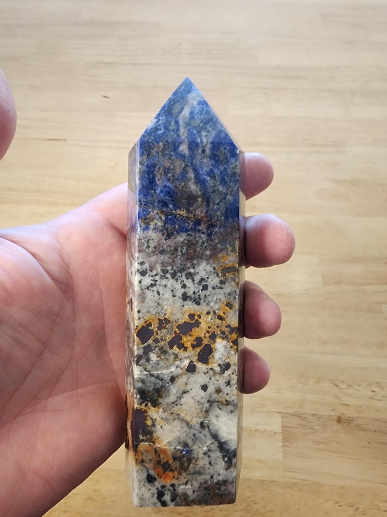 May include: A rectangular crystal point with a flat base and a pointed top. The crystal features a mix of colours, including deep blue, white, grey, and orange, with a polished surface. The crystal is held in a hand.