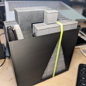 Könnte beinhalten: Ein schwarzer, 3D-gedruckter Schreibtisch-Organizer mit Fächern für Stifte und kleine graue Behälter. Ein gelbes Gummiband sichert die Behälter. Ein schwarzer und ein weißer Sharpie-Stift sind sichtbar.