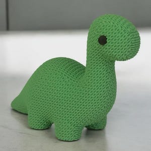 Pode incluir: Figura de dinossauro verde com textura de malha. O dinossauro tem um pescoço longo, pernas pequenas e uma cauda. Tem um olho escuro na lateral da cabeça. A figura é feita de um material macio.