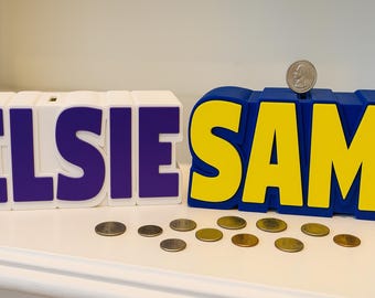 May include: Two personalized money banks, one white with purple letters spelling "ELSIE" and the other blue with yellow letters spelling "SAM". A coin sits on top of the "SAM" bank, with loose change scattered in front.