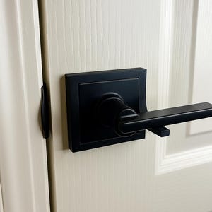 Peut inclure: Gros plan d'une poignée de porte noire avec une plaque arrière carrée et un levier. La poignée est montée sur une porte blanche avec des panneaux verticaux. La porte est légèrement entrouverte, révélant un loquet de porte noir sur le cadre.