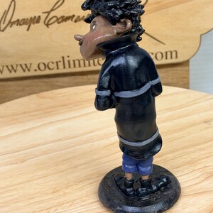 May include: A handcrafted figurine of a person in a black coat with gray accents and blue shorts. The figure has dark skin and curly black hair. The figurine is standing on a dark, round base.
