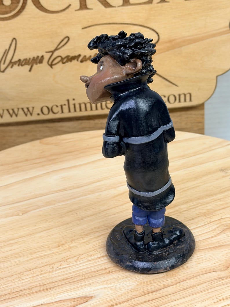May include: A handcrafted figurine of a person in a black coat with gray accents and blue shorts. The figure has dark skin and curly black hair. The figurine is standing on a dark, round base.