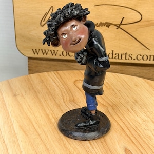 May include: A handcrafted figurine of a person in a black coat, blue pants, and black shoes, standing on a black circular base. The figure has a light skin tone, curly black hair, and is looking down. The background includes a wooden surface and text.