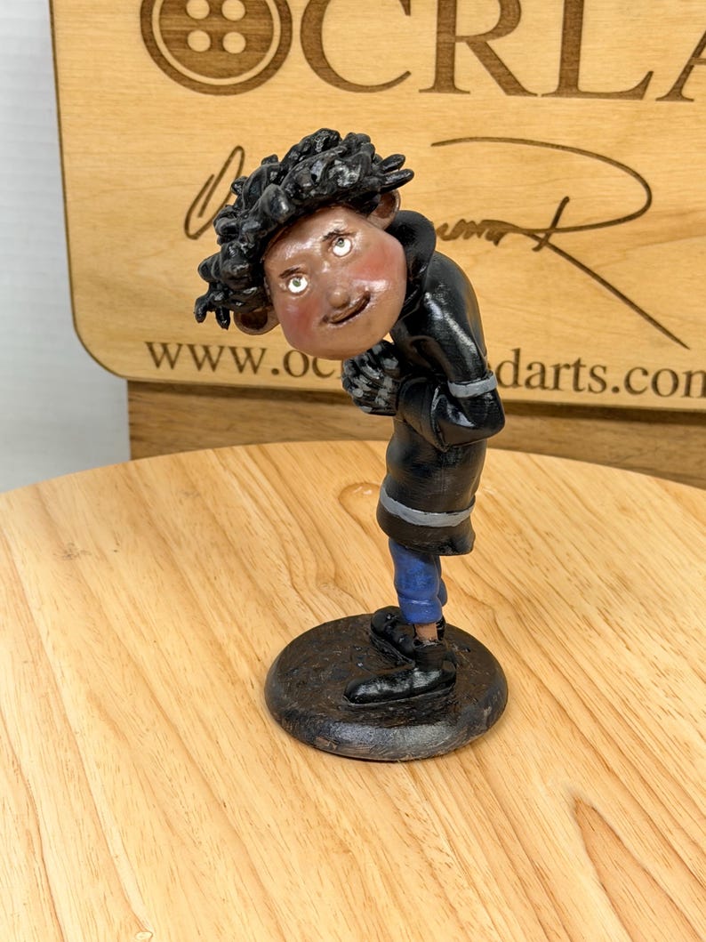 May include: A handcrafted figurine of a person in a black coat, blue pants, and black shoes, standing on a black circular base. The figure has a light skin tone, curly black hair, and is looking down. The background includes a wooden surface and text.