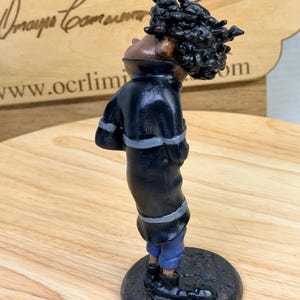 May include: A handcrafted figurine of a person in a black coat with gray trim and blue pants. The figure has a dark complexion and a large, textured hairstyle. The figurine stands on a dark, circular base.